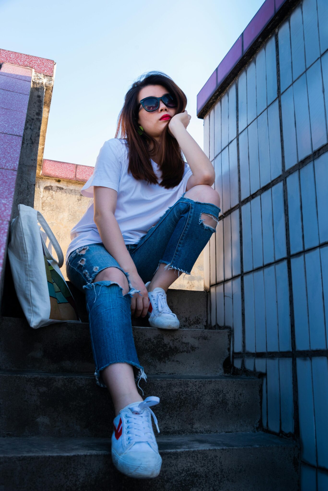 A stylish woman in sunglasses and casual attire sits thoughtfully on outdoor stairs.