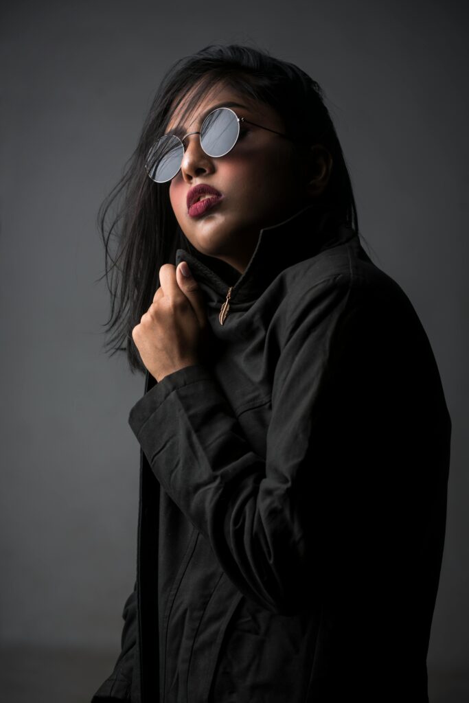 Stylish portrait of a woman posing in a studio with dark fashion attire and round sunglasses.